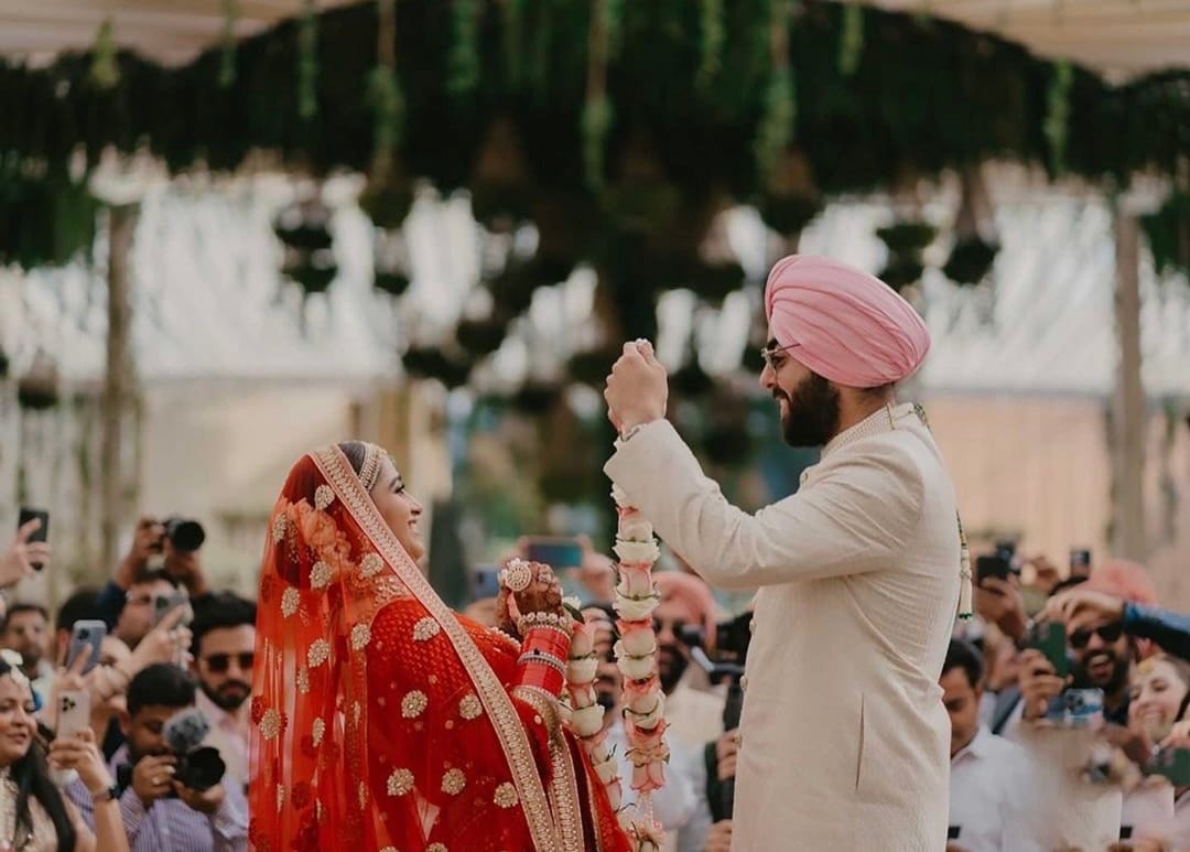 The groom lifts the varmaala above the bride while a ring of family films it on their phones