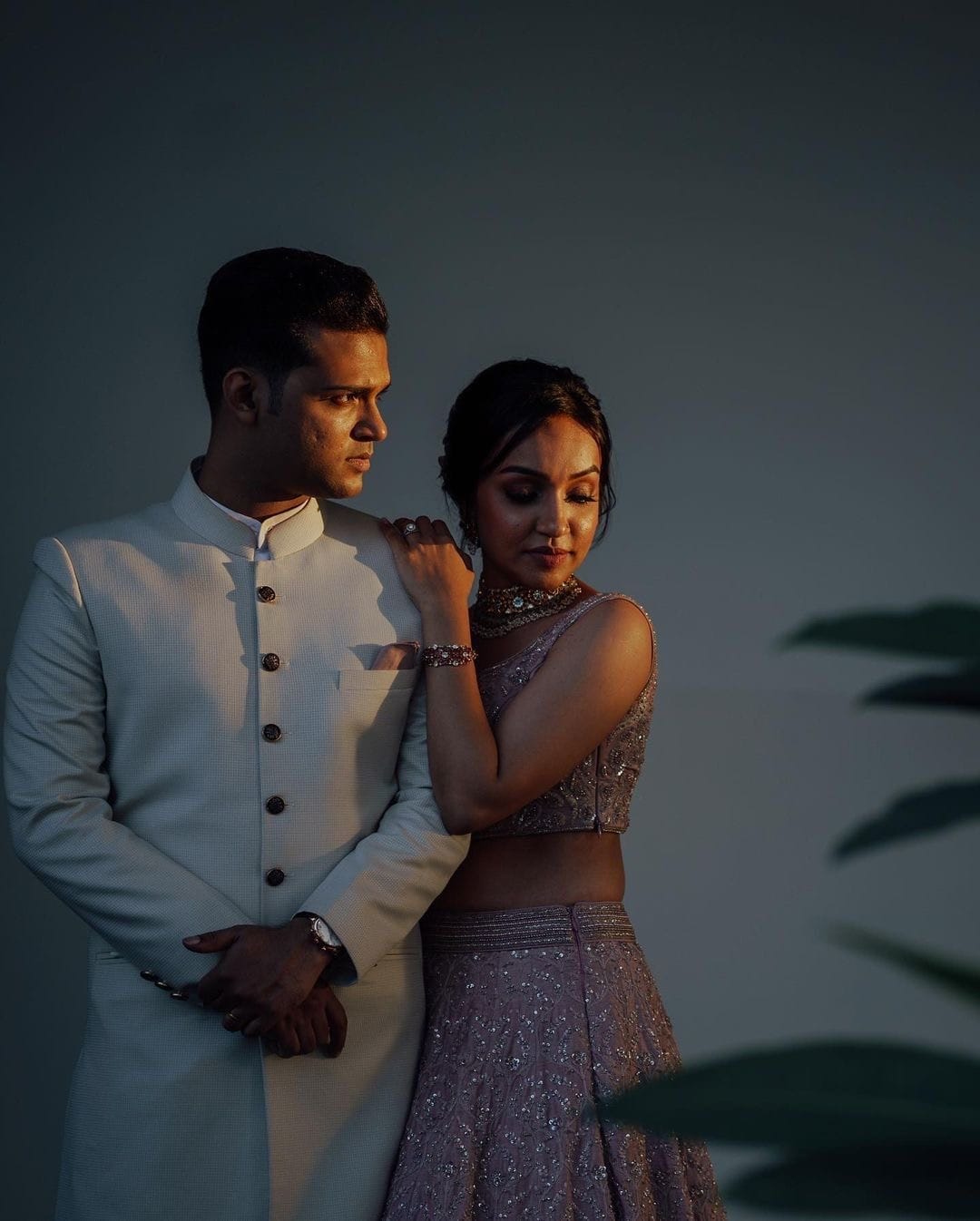 Hard evening light across a plain wall, she rests a hand on his shoulder in a lilac lehenga