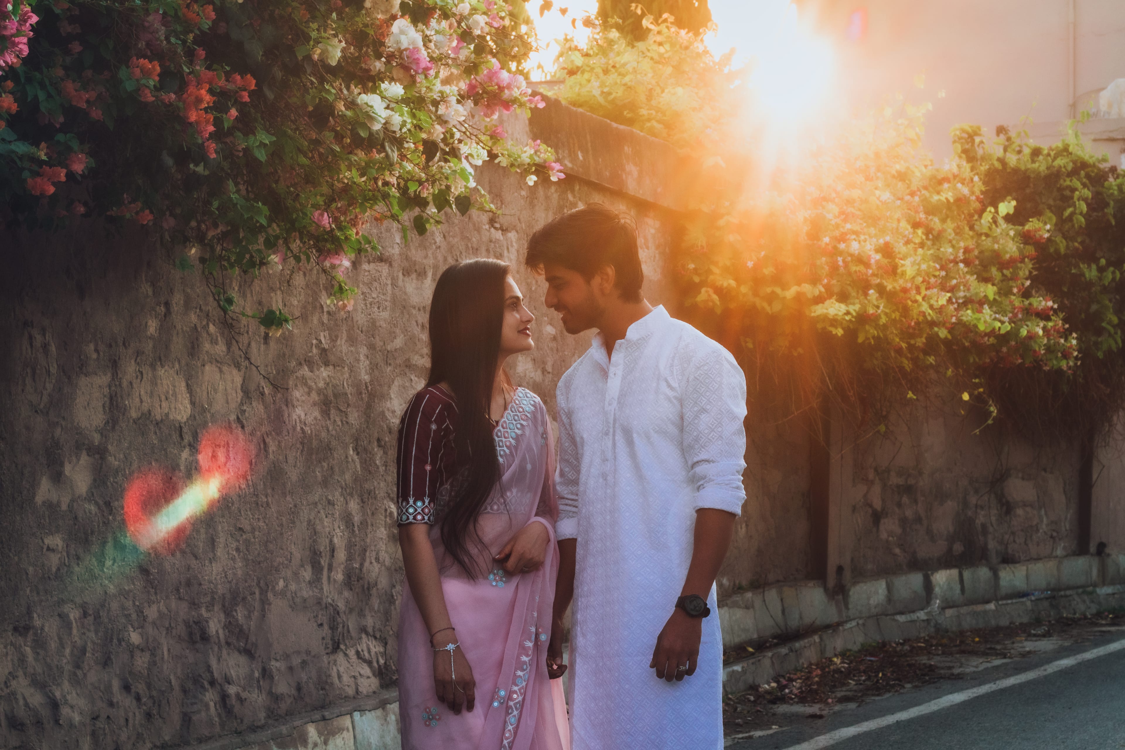 Couple facing each other on a lane at sunset, bougainvillea spilling over a stone wall, the sun flaring behind them