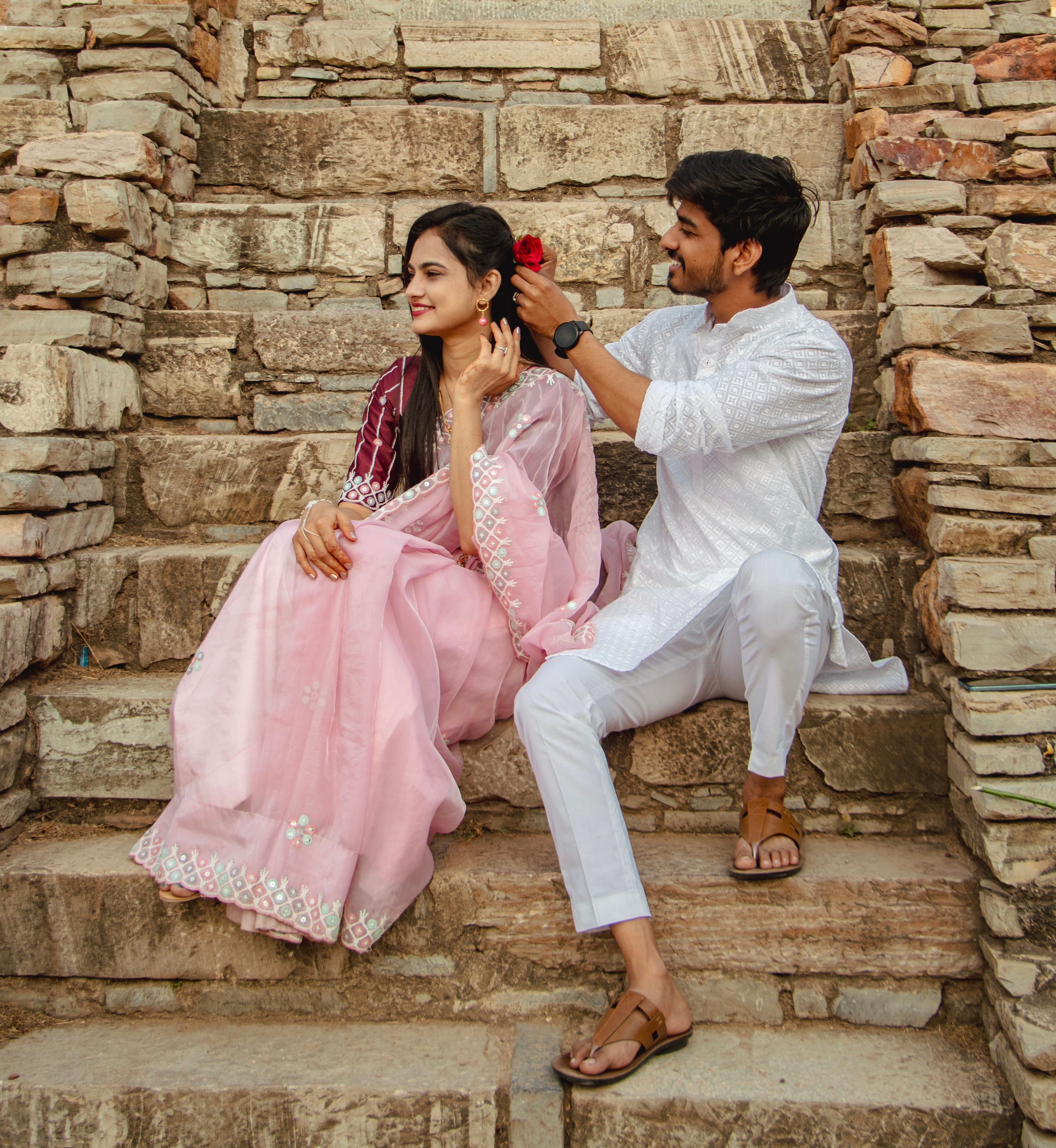 He tucks a red rose into her hair as they sit on the worn stone steps of a fort