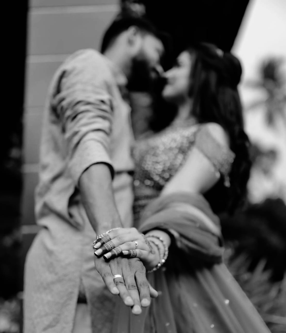 Their linked hands and rings held towards the camera, the couple kissing out of focus behind