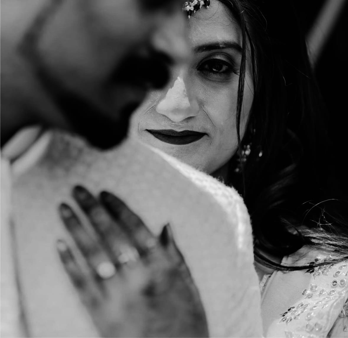 Her eye meets the camera over his shoulder, a mehndi covered hand resting on his back