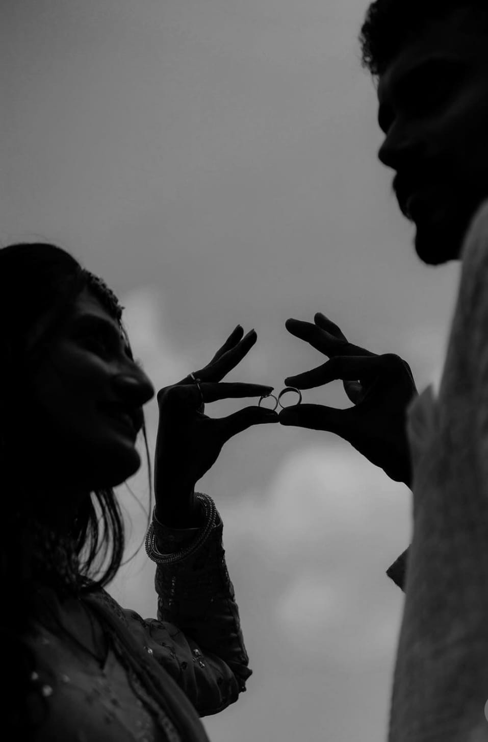 Silhouette of the couple holding two rings pinched between their fingertips against a pale sky