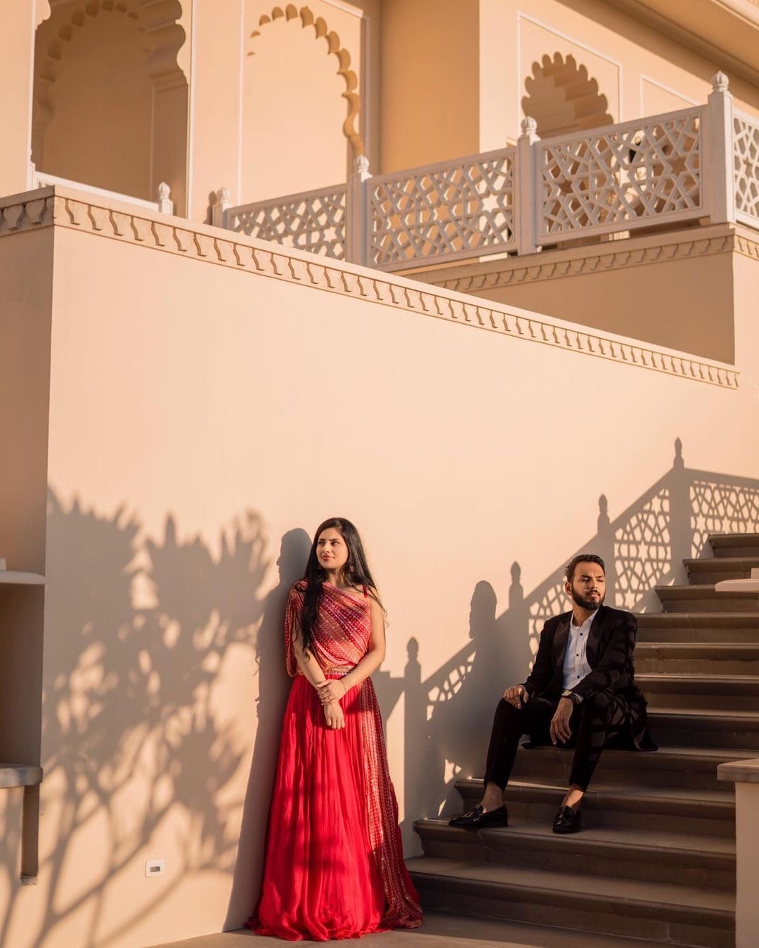 Jaali shadows fall across a pink palace wall, she leans against it and he sits on the steps