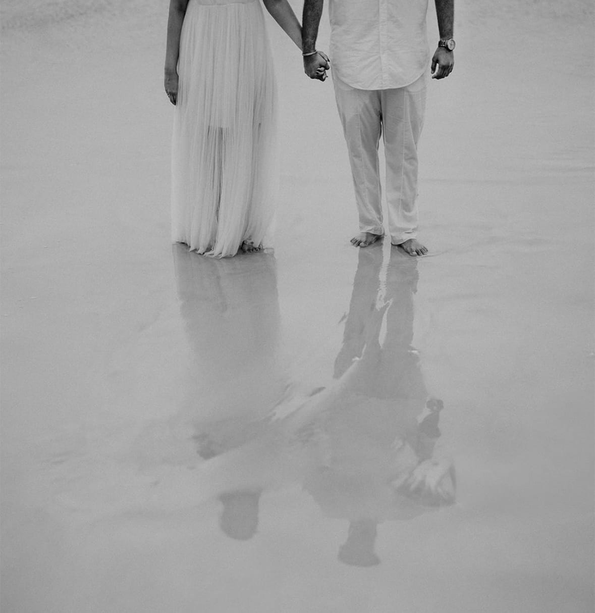 Two pairs of bare feet and a pair of held hands reflected in wet sand