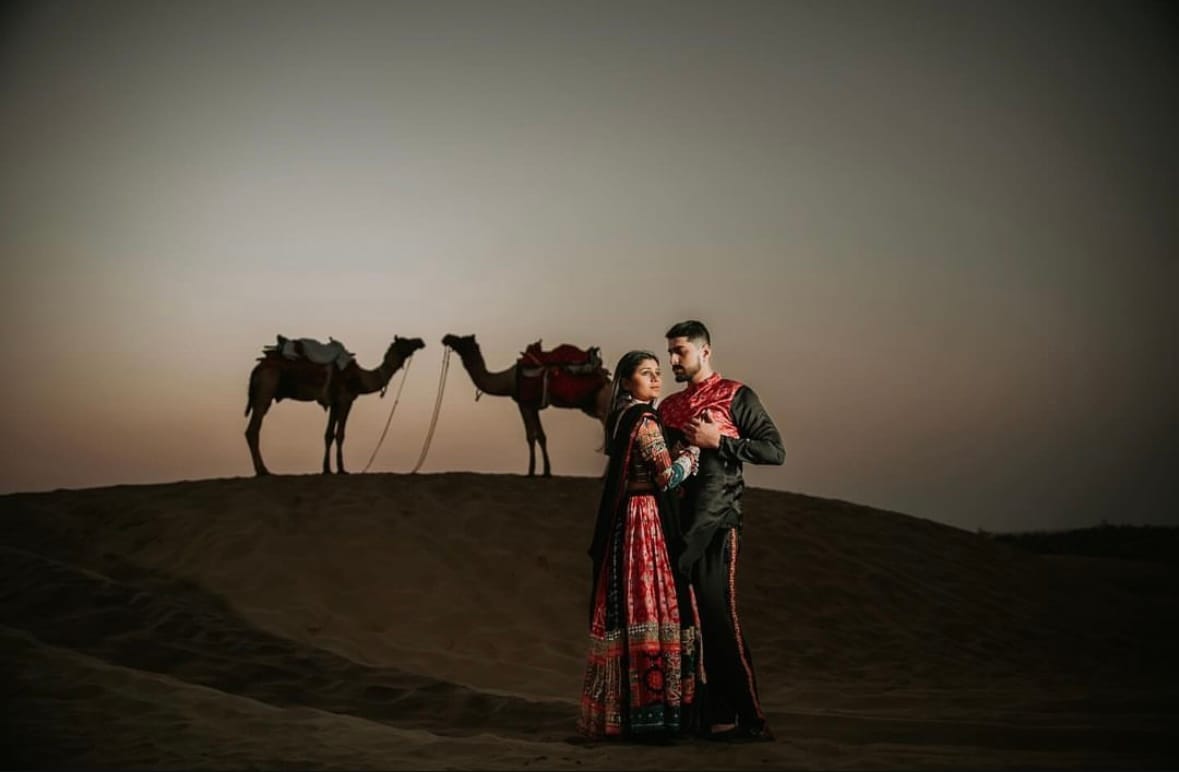 Couple on a dune at dusk in the Thar desert, two saddled camels on the ridge behind them