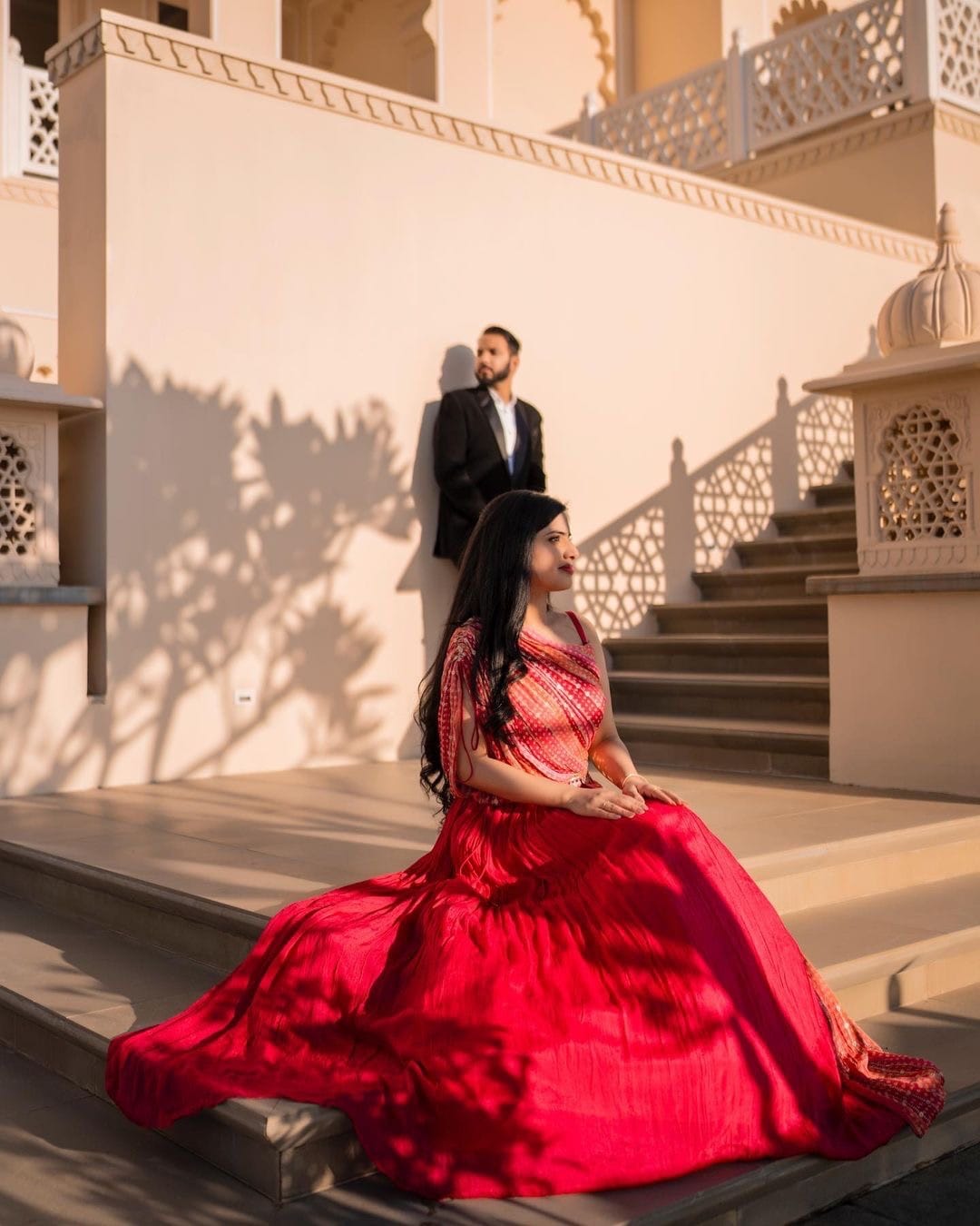 She sits on palace steps with her red skirt spread around her, he leans on the wall behind