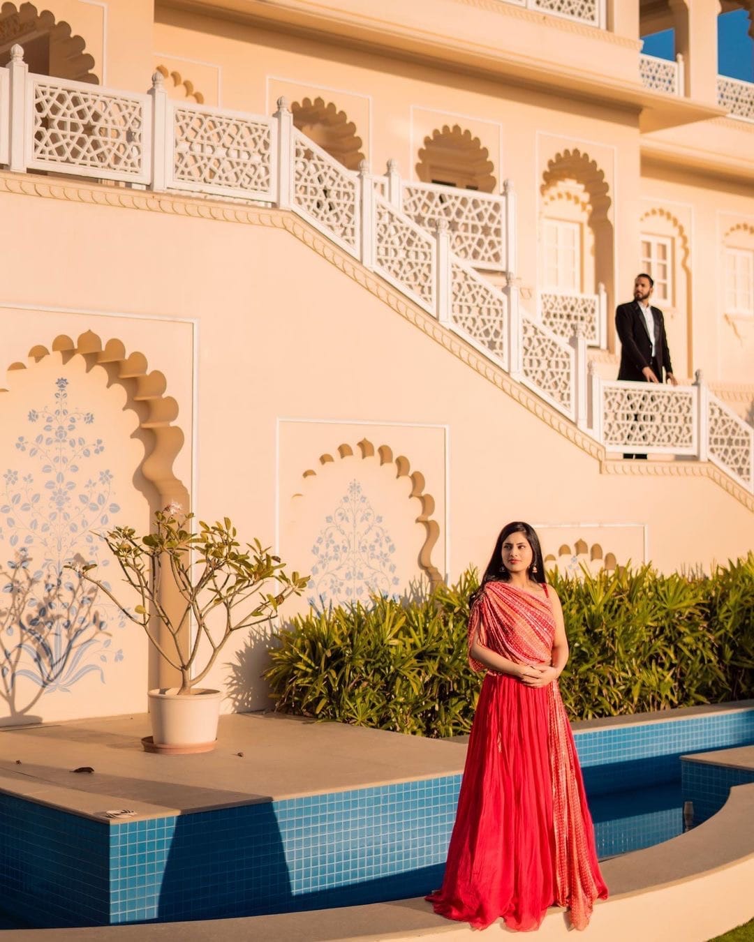 She stands beside a palace pool in a red gown while he watches from the staircase above