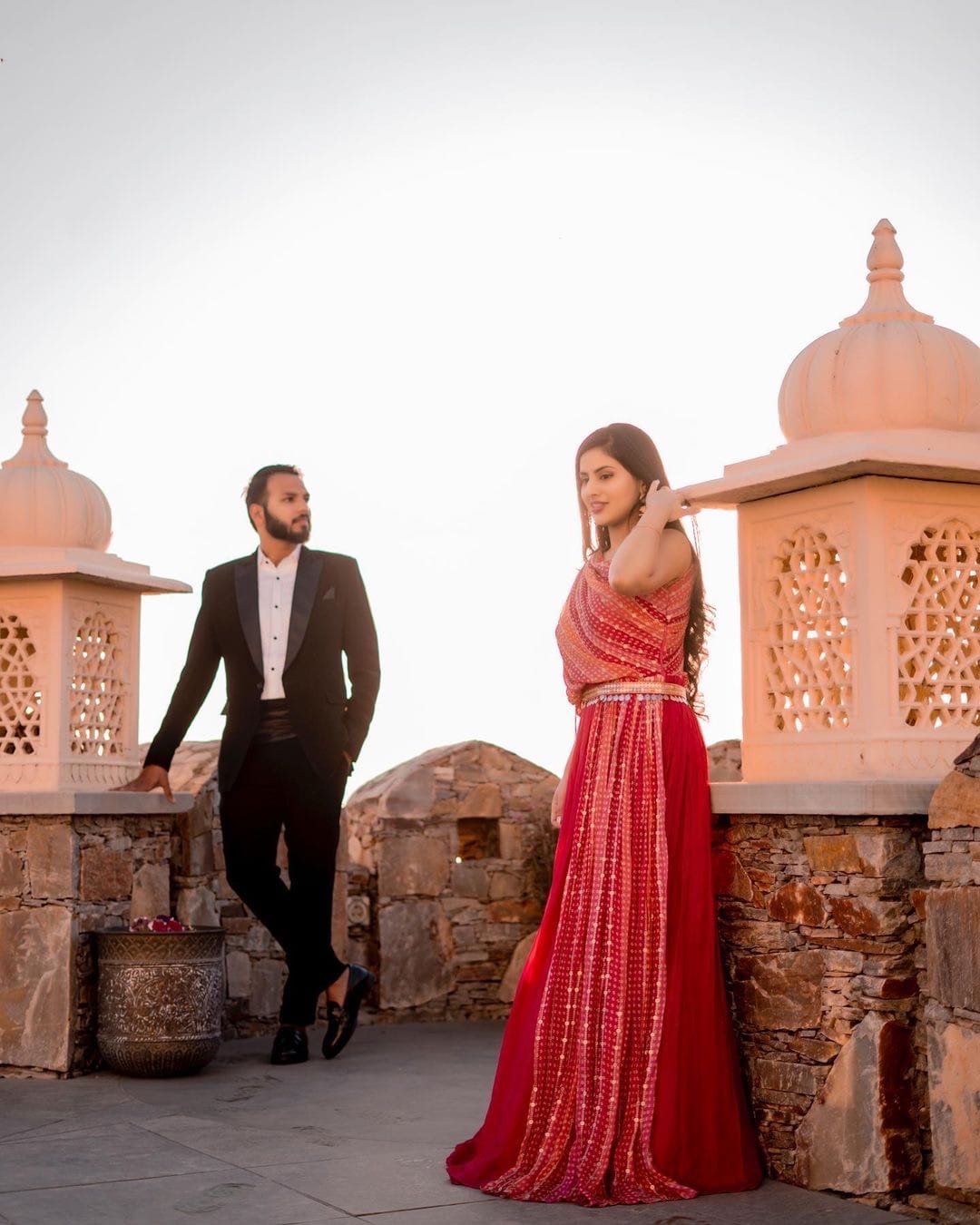She turns towards the camera on a Rajasthan fort wall, he leans against a chhatri behind her