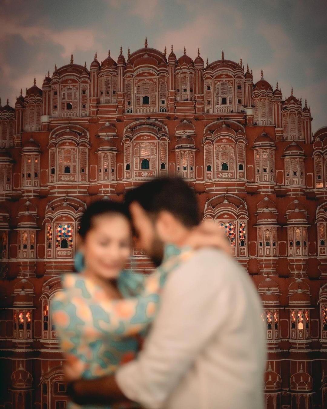 Hawa Mahal sharp at dusk while the couple embrace, deliberately out of focus in the foreground
