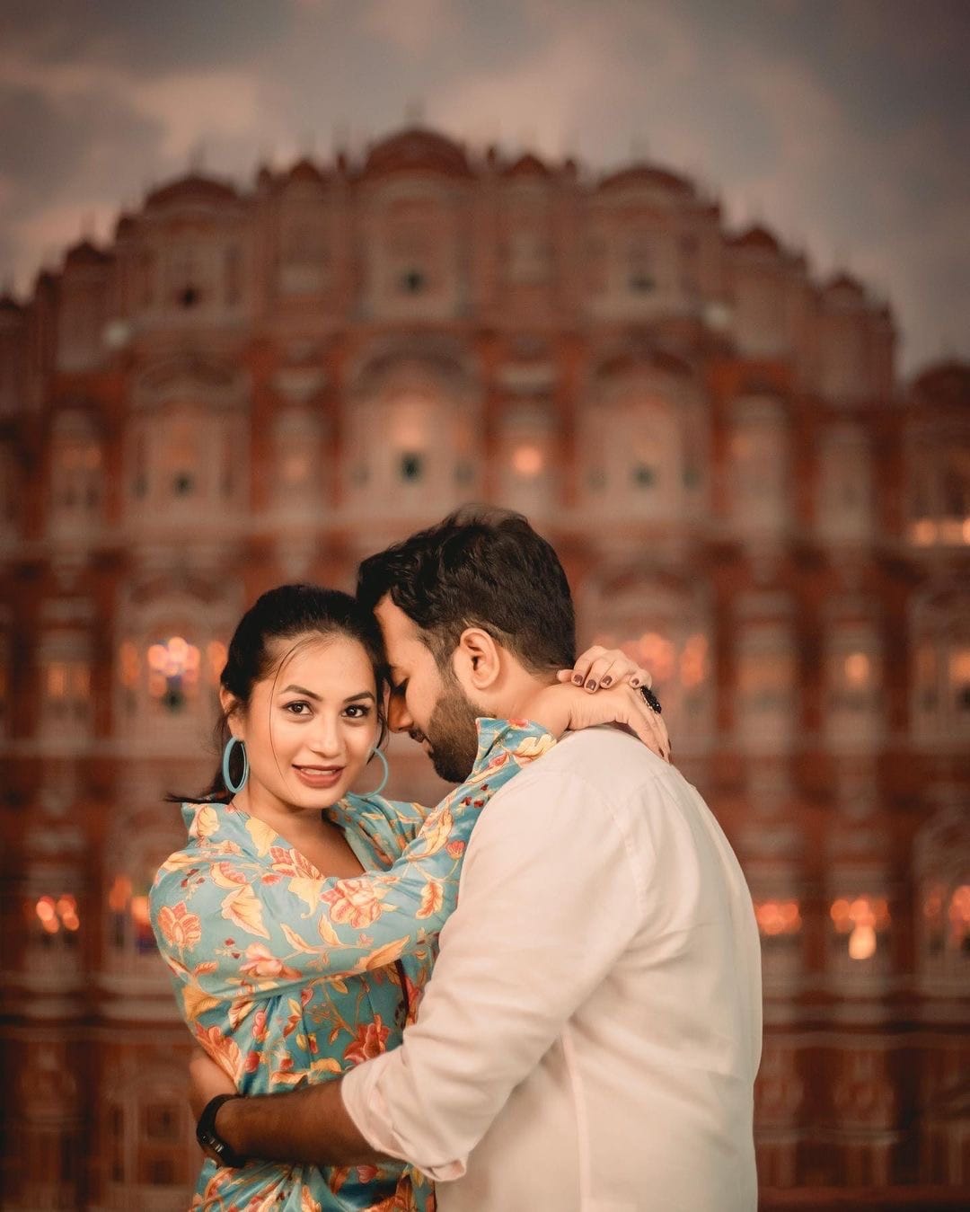 She looks back over his shoulder at the camera, arms around his neck, Hawa Mahal glowing behind them