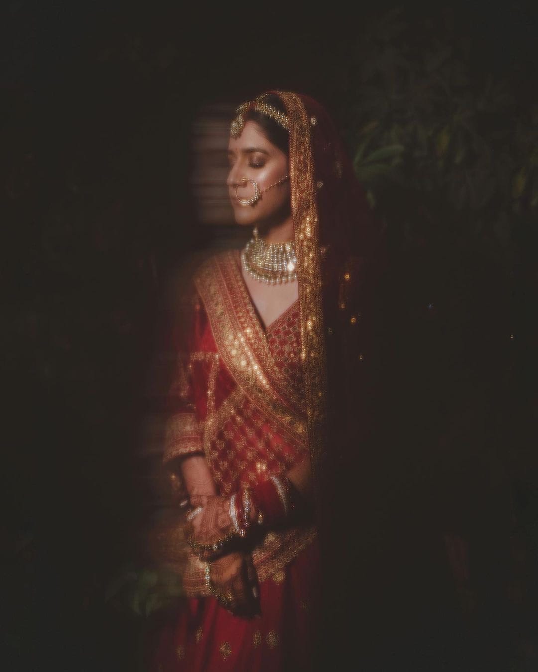Bride in red and gold standing in near darkness, eyes lowered, a gold zari veil across her shoulder