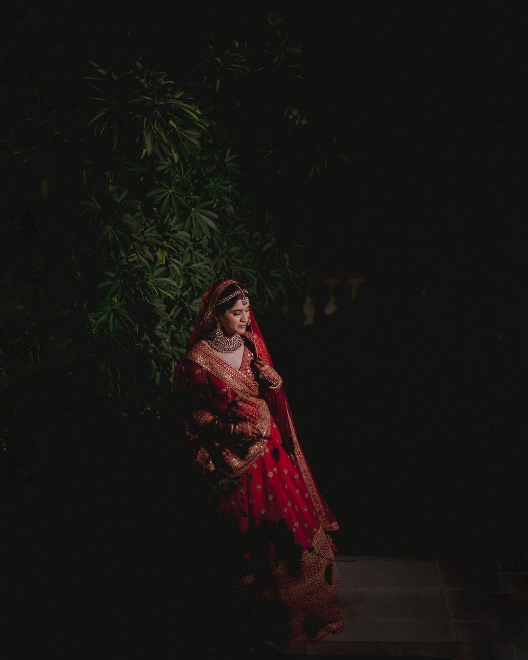 Bride alone in red and gold against a dark tree, lit from one side