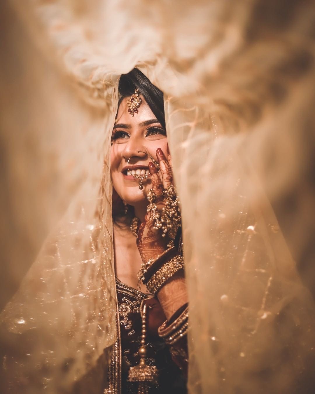 Bride framed through a gap in a gold veil, one hand raised, smiling past the camera