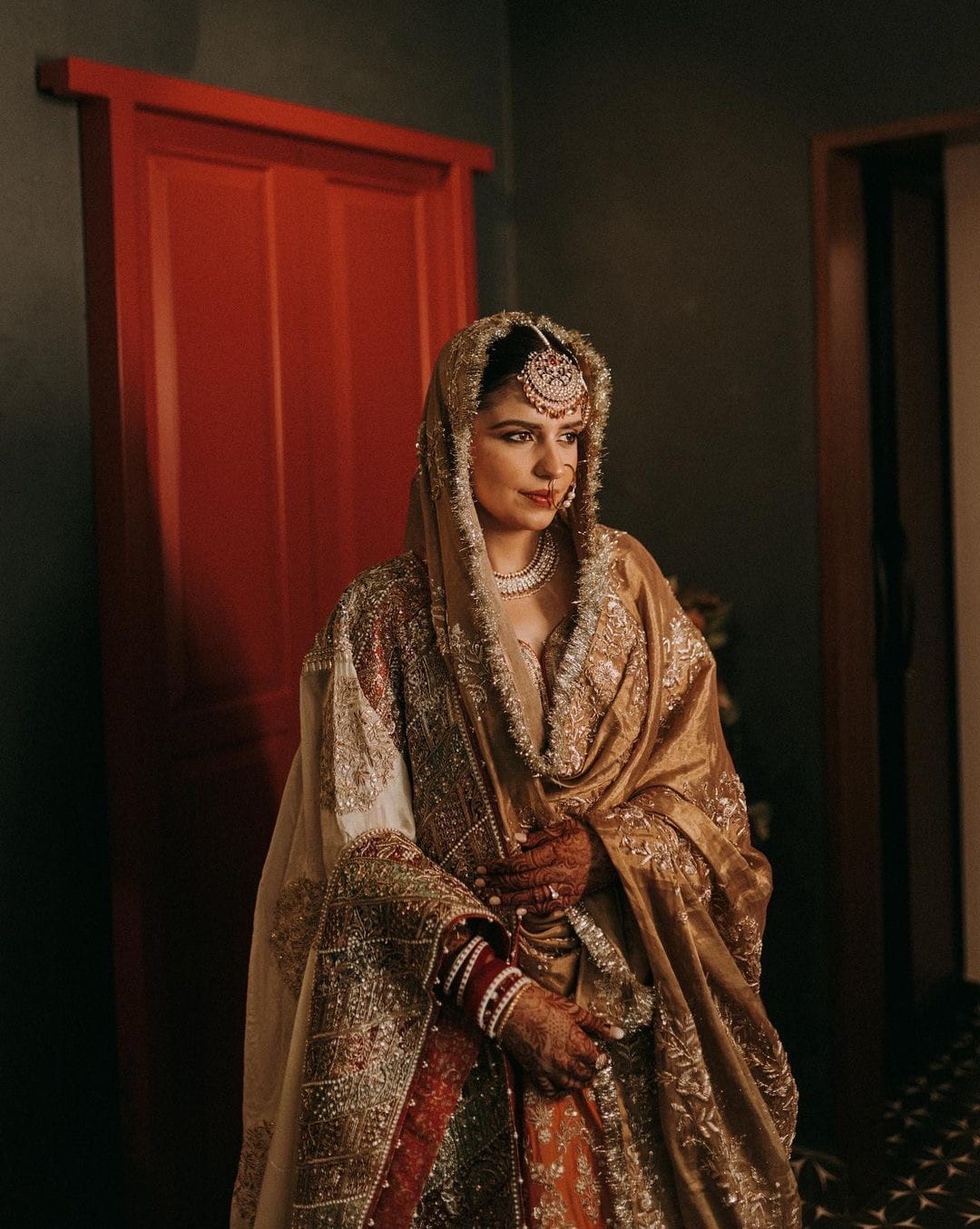 Bride in a gold tissue dupatta beside a red door, half in shadow