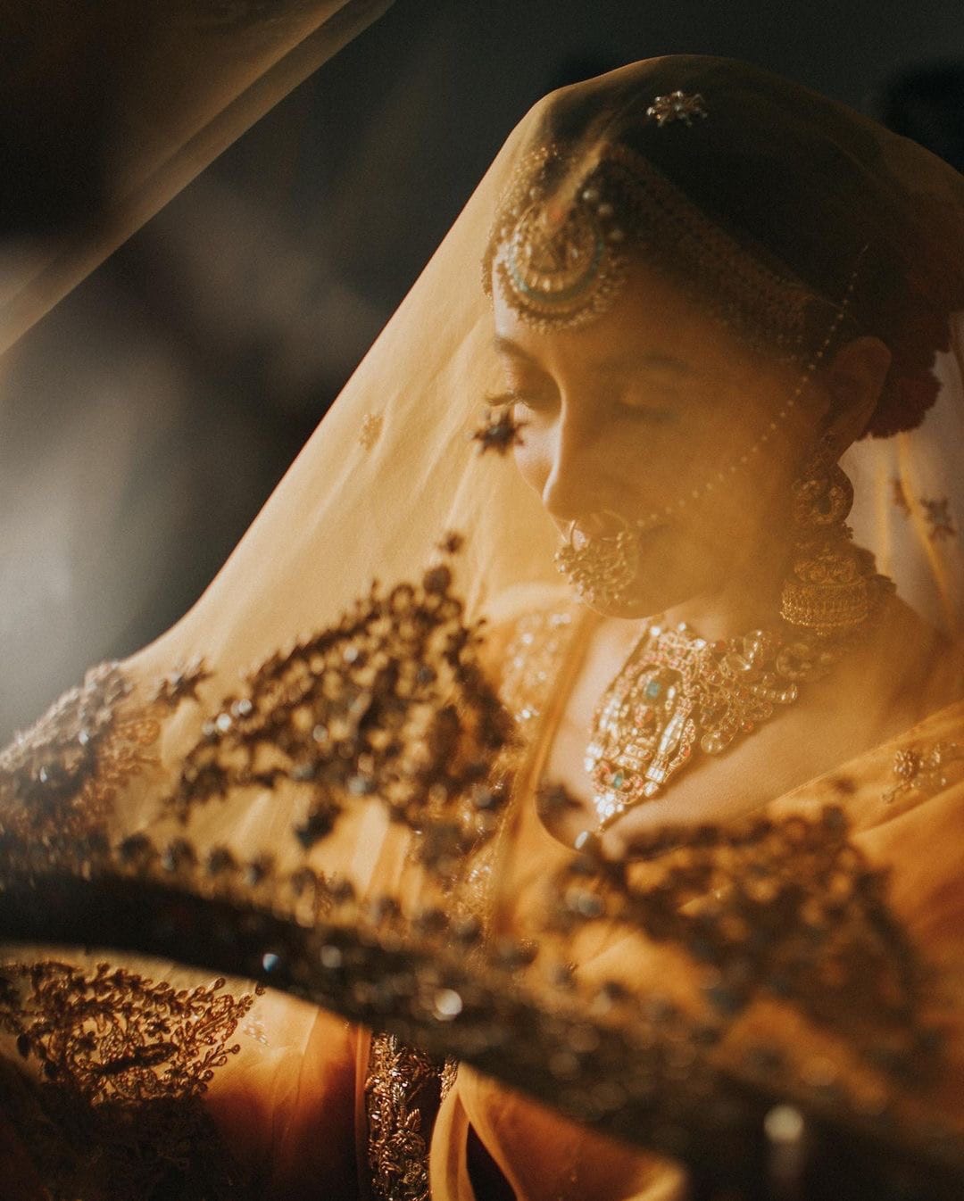 Bride under a gold veil in tungsten light, jhumkas and a layered kundan necklace catching the glow