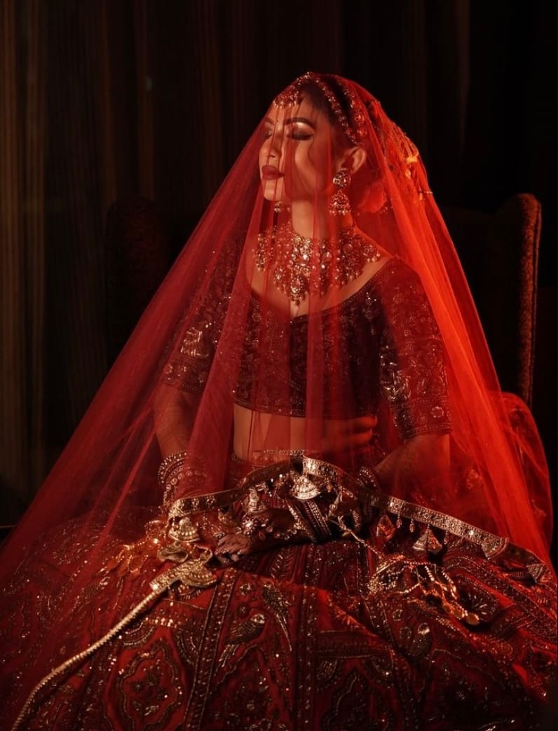 Bride seated under a red veil, a gold kamarbandh across her lap, lit by a single warm source