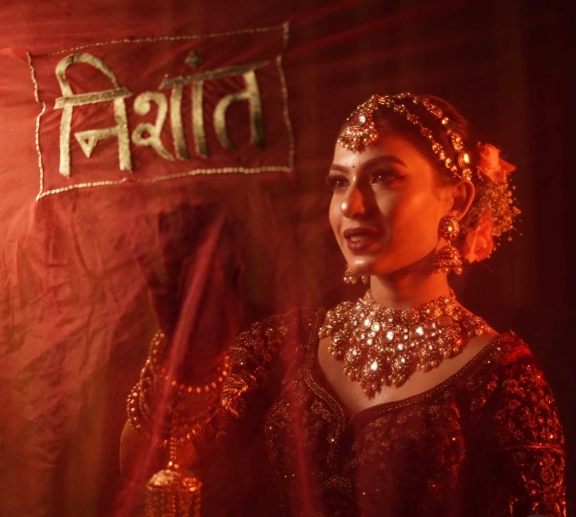 Bride beside a red drape with a name embroidered in Devanagari, warm haze across the frame