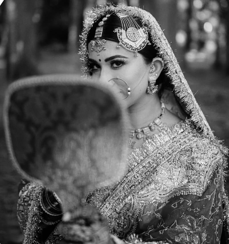 Bride in black and white, half hidden behind a hand fan, a heavy maang tikka across her forehead