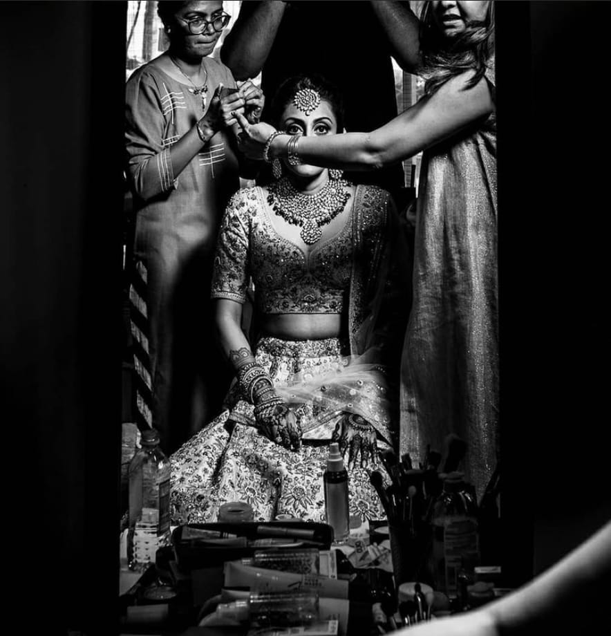 Bride seated at a makeup table in black and white while two pairs of hands finish her hair