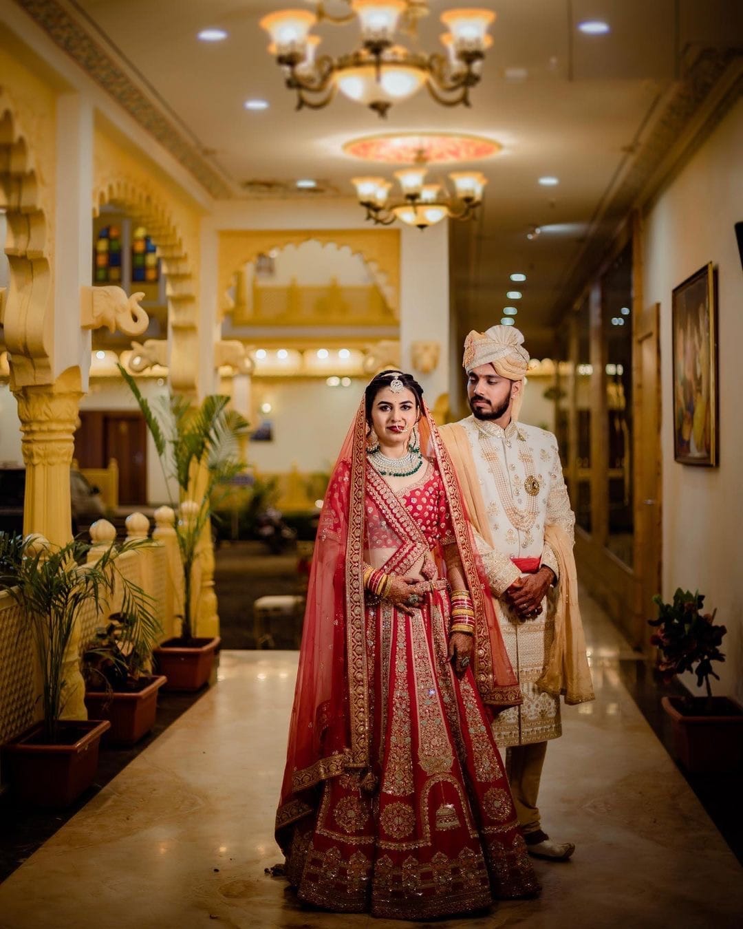 Couple standing in a yellow sandstone corridor, her red lehenga trailing on the marble
