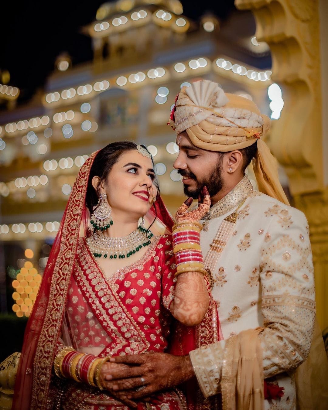 Bride reaches up to the groom's chin, a lit haveli facade blurred behind them
